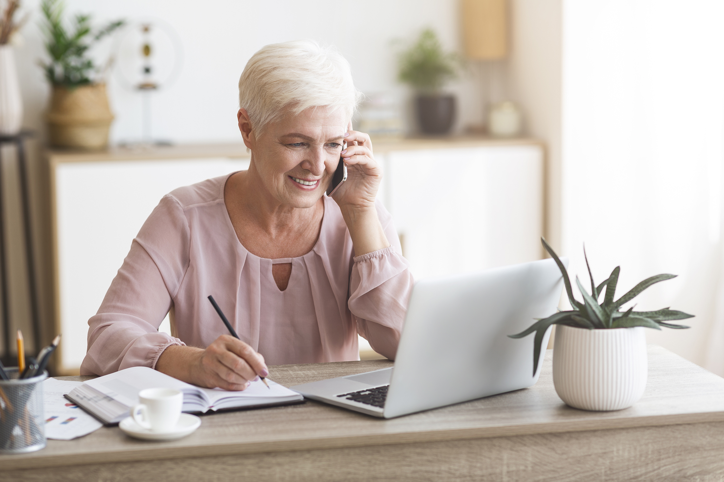 Senior business woman working from home, discussing project with client, making notes in front of laptop, panorama with empty space